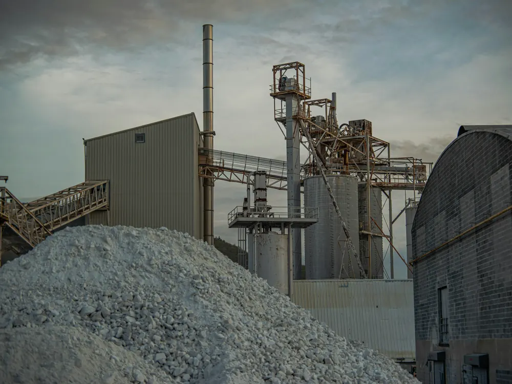 Stockpile of crushed pale stone in front of a processing plant with an inclined conveyor, steel silos, an elevator tower and a chimney at dusk