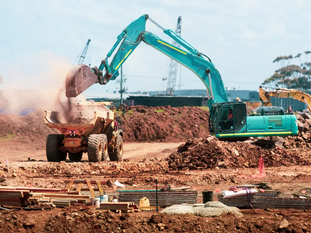 Teal tracked excavator tipping soil into an articulated dump truck on a red-earth construction site, with cranes in the background