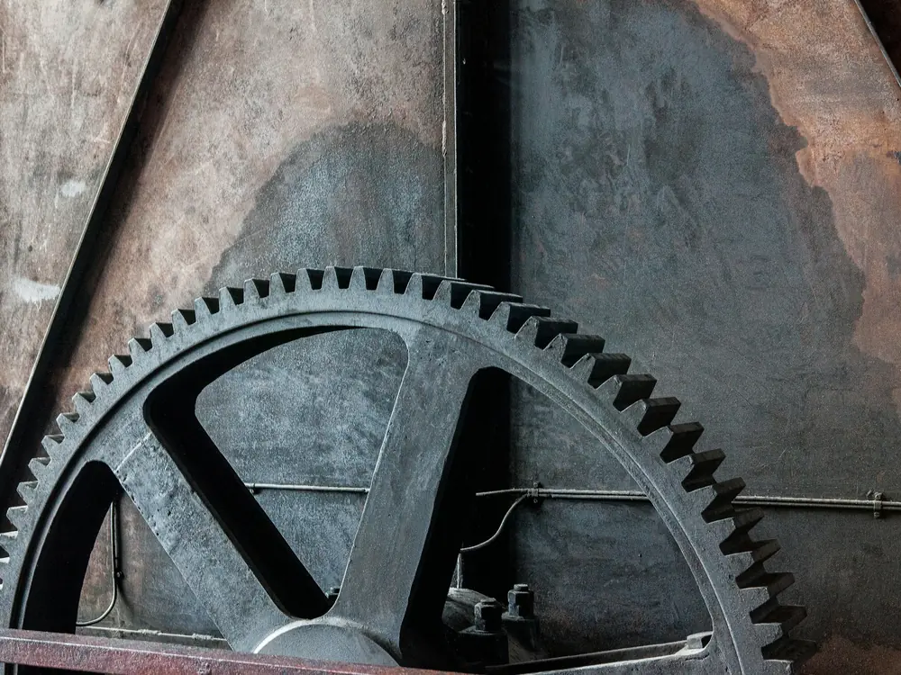 Large cast spur gear wheel with heavy spokes set against weathered, riveted steel plate
