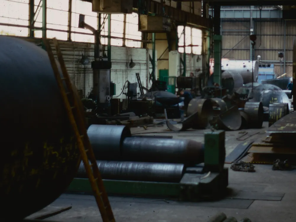 Heavy fabrication shop with a plate-rolling machine in the foreground and rolled steel shells and plate laid out across the floor