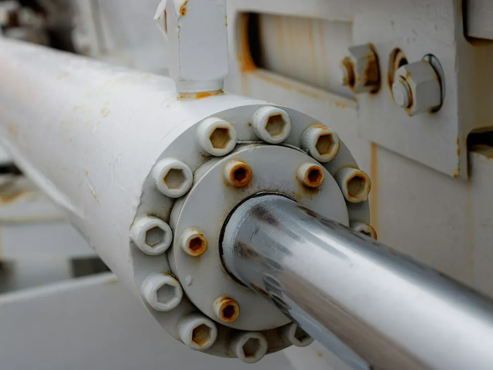 Close-up of a white-painted hydraulic cylinder end cap with socket-head bolts and a polished chrome piston rod
