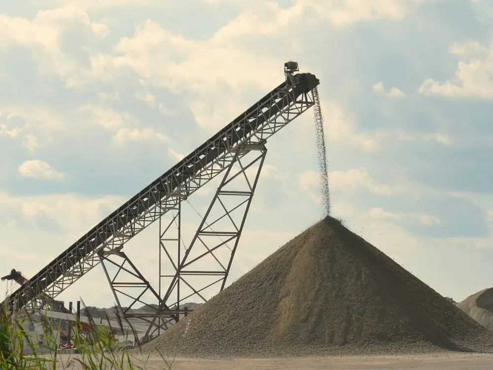 Inclined lattice-frame conveyor discharging aggregate onto a conical stockpile under a cloudy sky