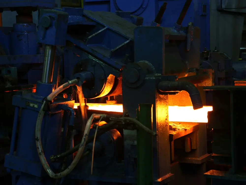 Glowing hot steel billet passing through a blue rolling-mill stand fitted with cooling-water hoses